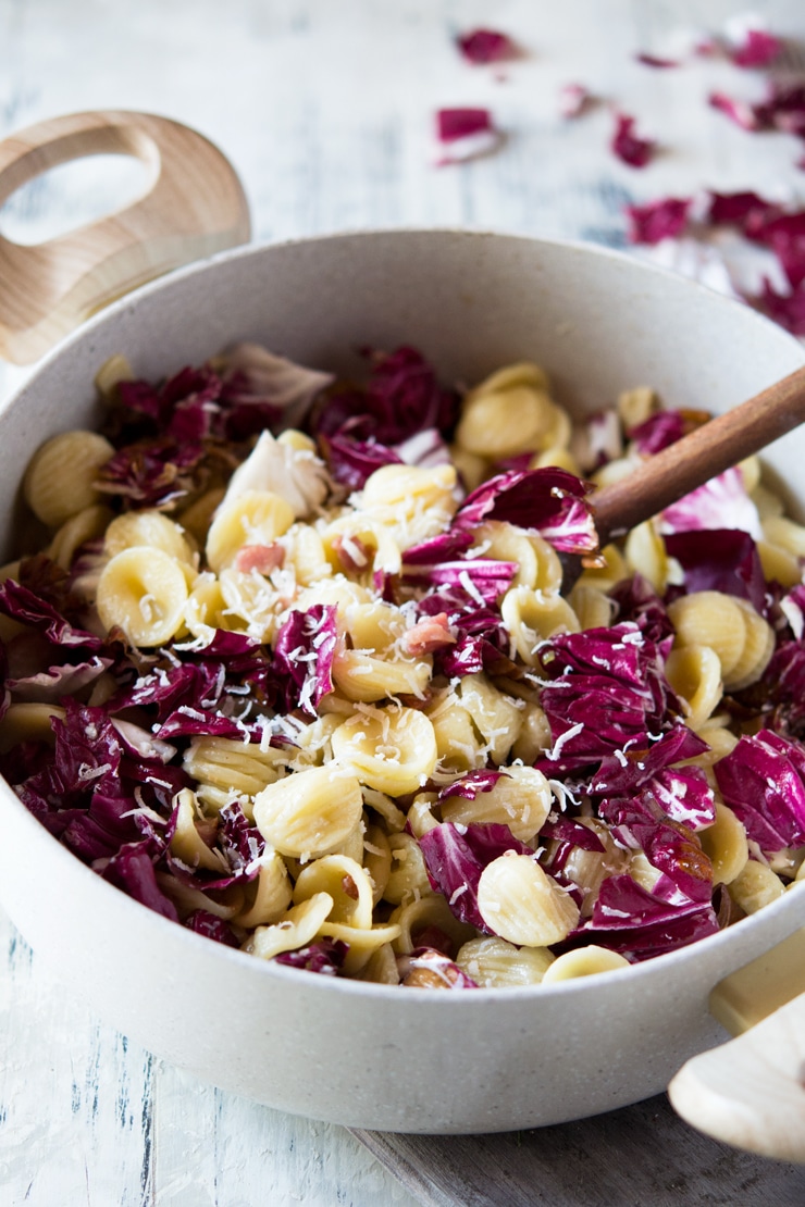 A close up of orecchiette pasta with pancetta and radicchio with a wooden spoon in a large pan