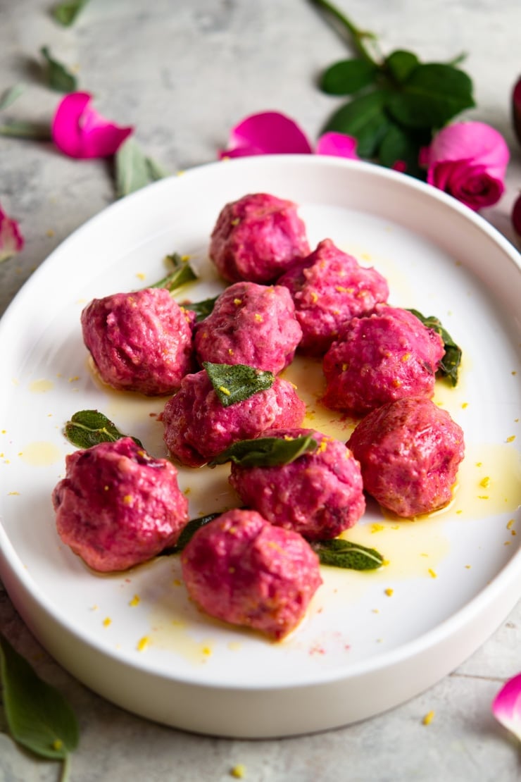 A close up of beetroot ricotta gnudi on a white plate with sage and butter