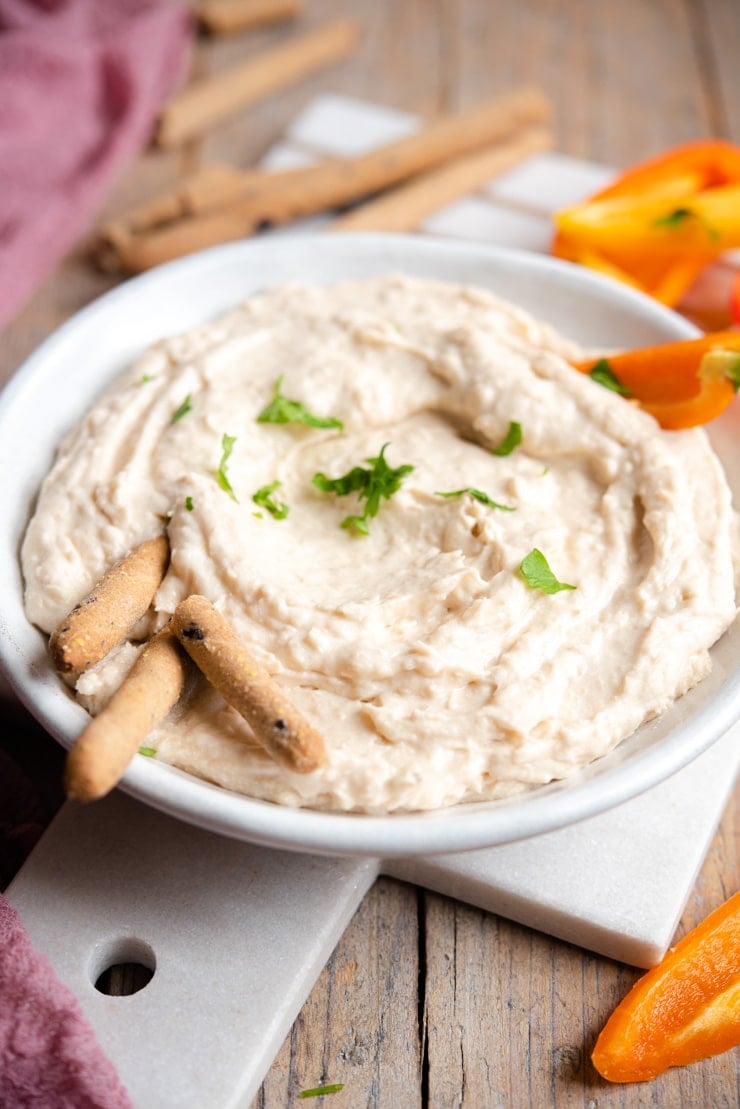 A side shot of a white bean dip with breadsticks and bell peppers around it