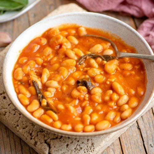 A bowl of cannellini bean in tomato sauce with sage sitting on a wooden surface