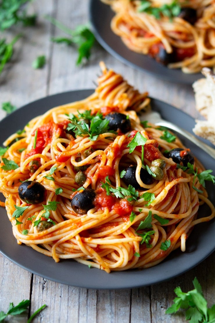 A close up of spaghetti on a plate with puttanesca sauce sitting on a wooden surface