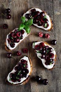 Whipped ricotta toast recipe with deep red, fresh cherries tossed in balsamic vinegar and scattered with some chopped basil leaves. Easy Italian appetizers at inside the rustic kitchen.