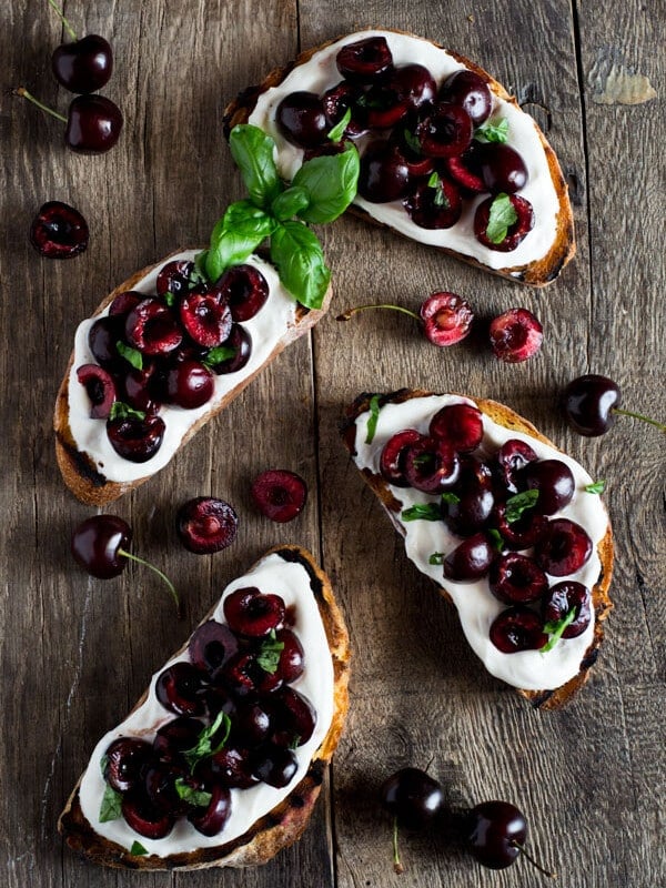 Whipped ricotta toast recipe with deep red, fresh cherries tossed in balsamic vinegar and scattered with some chopped basil leaves. Easy Italian appetizers at inside the rustic kitchen.