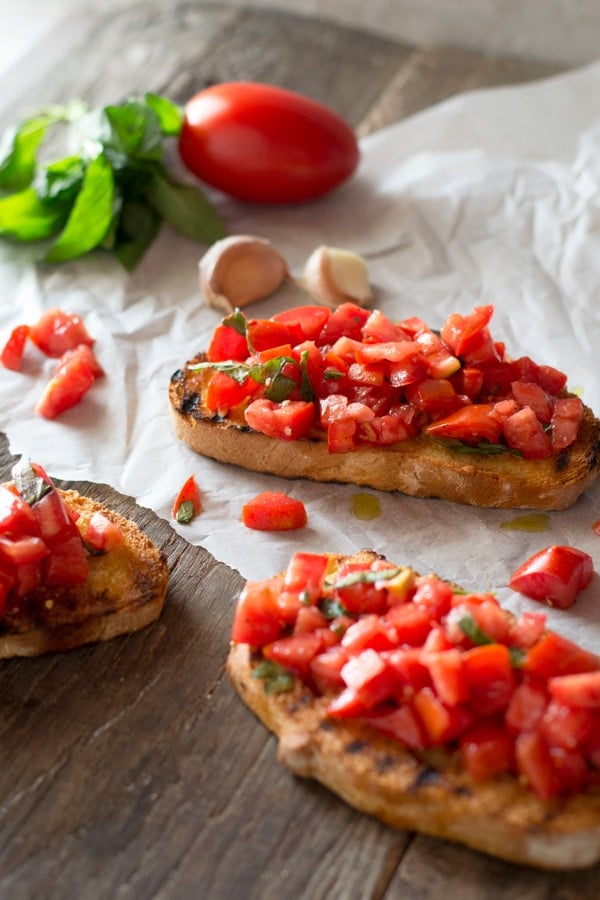 Tomato bruschetta sitting on a wooden surface with garlic, tomato and basil in the back ground