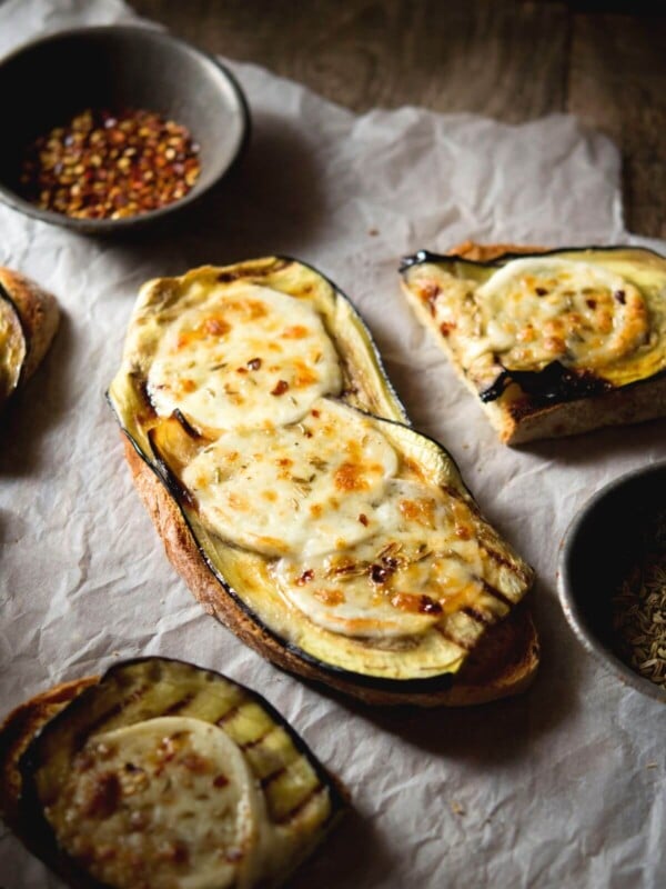 A slice of eggplant bruschetta in a rustic setting topped with mozzarella, red pepper flakes and fennel seeds.