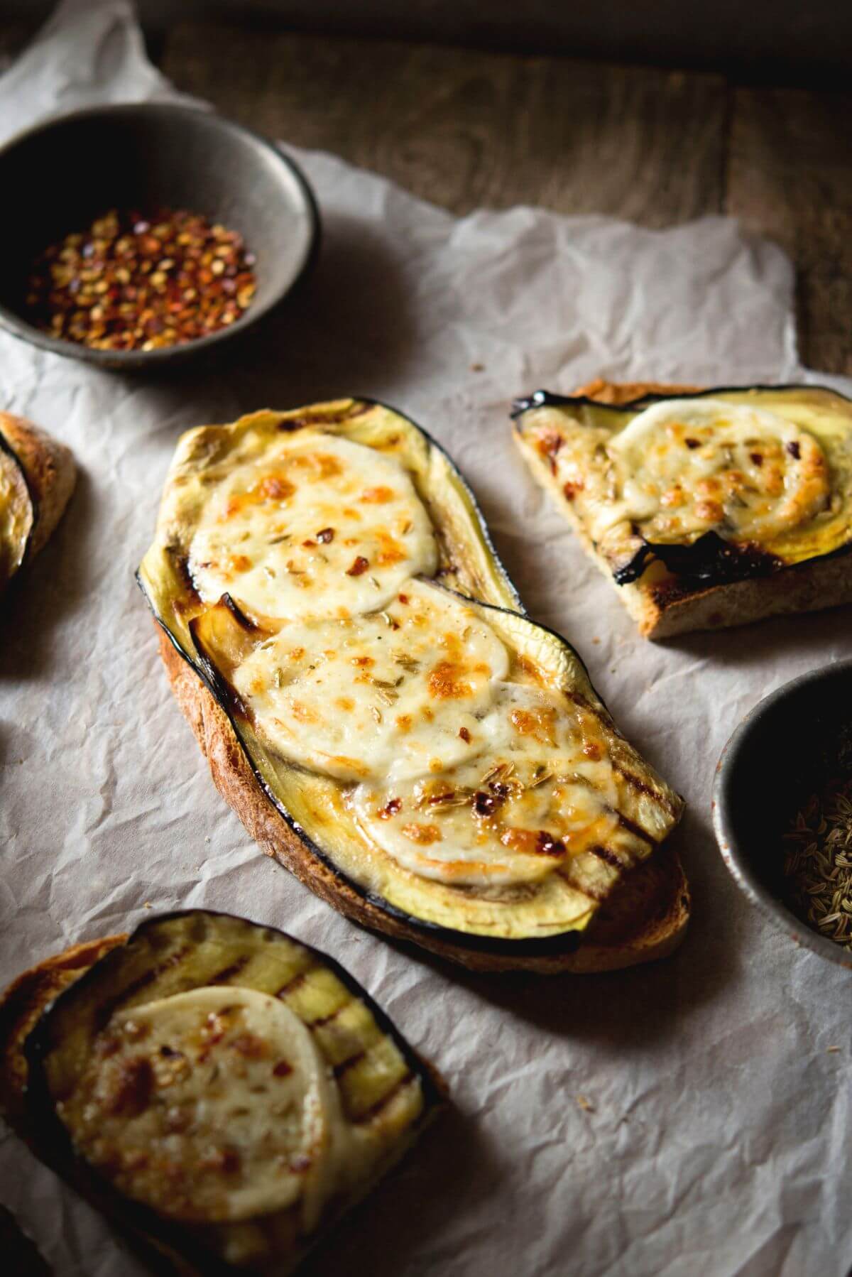 A slice of eggplant bruschetta in a rustic setting topped with mozzarella, red pepper flakes and fennel seeds.