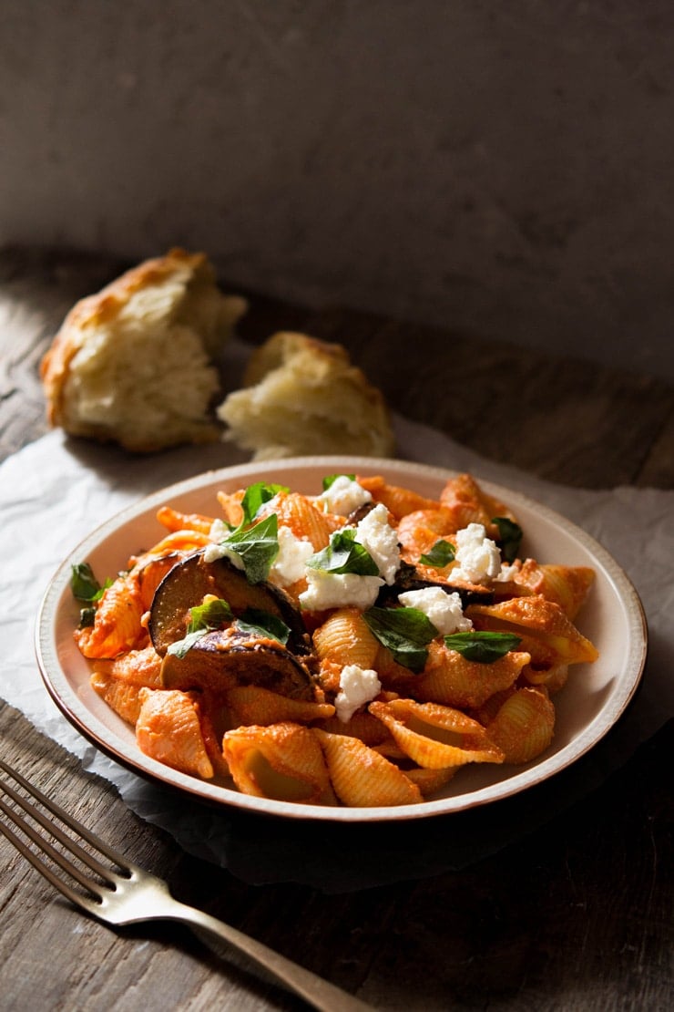 Eggplant pasta in tomato sauce in a bowl topped with ricotta cheese and basil torn crusty bread in the background