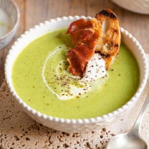 A close up of green pea soup with a slice of bread and prosciutto at the side