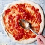 A hand spreading tomato sauce on a pizza base sitting on a marble surface.