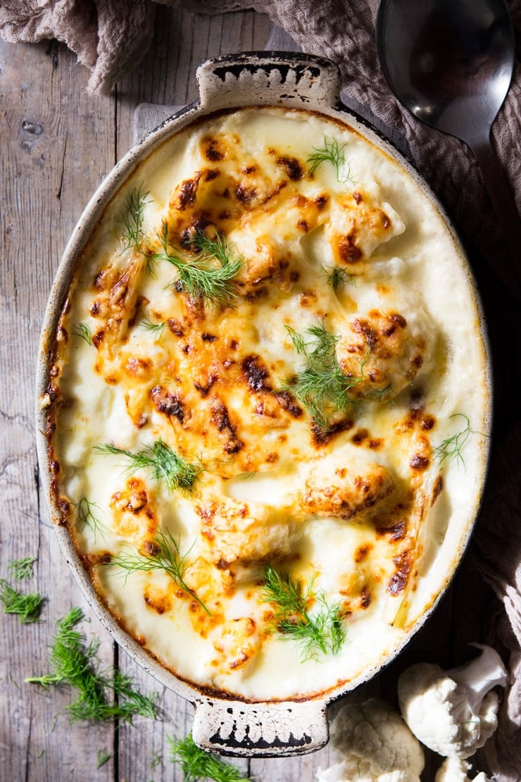 An overhead shot of baked cauliflower cheese in a oval baking dish on a wooden surface
