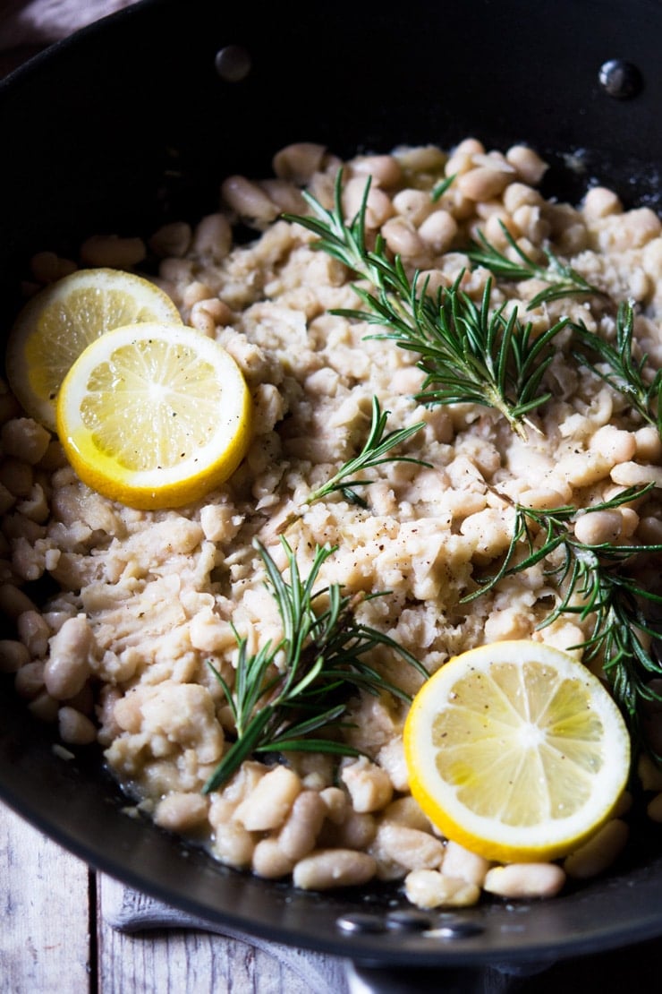 A close up of cannellini bean mash topped with sprigs of rosemary and slices of lemon in a large pan