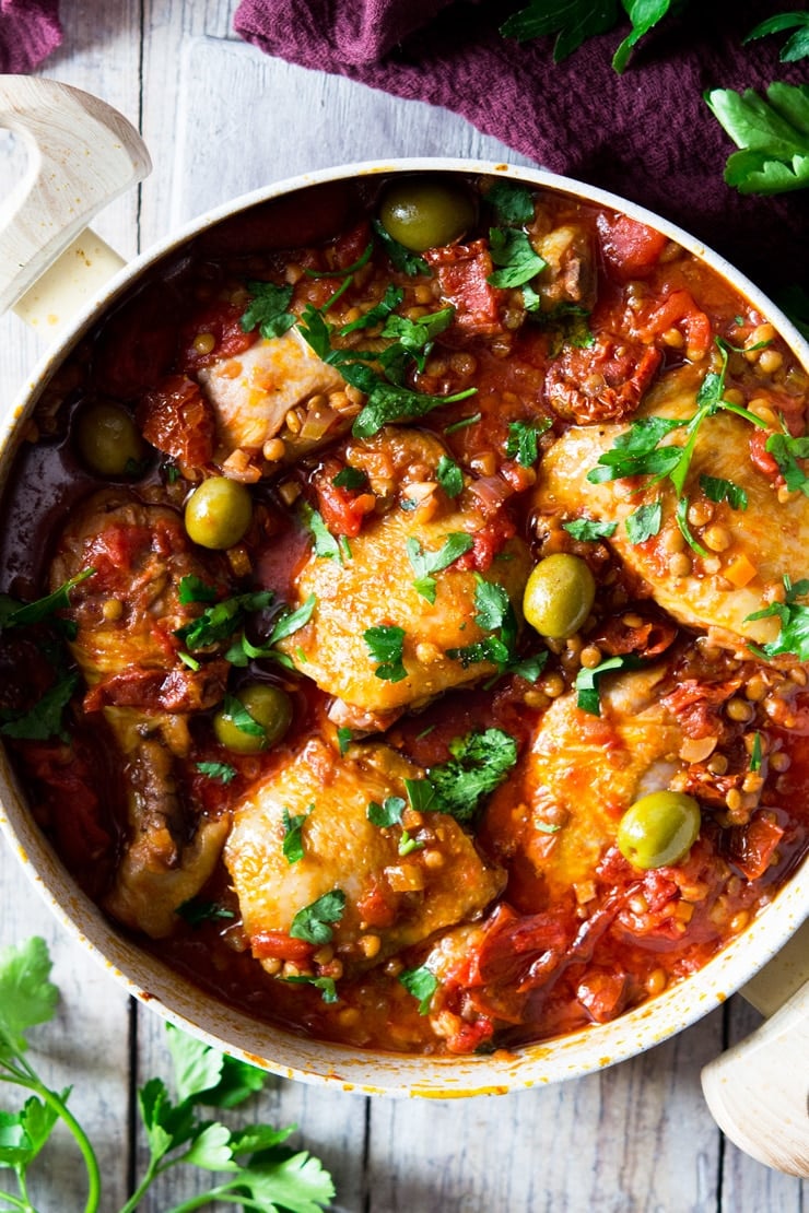 an overhead shot of braised chicken with olives, tomatoes and lentils in a pot sitting on a wooden surface