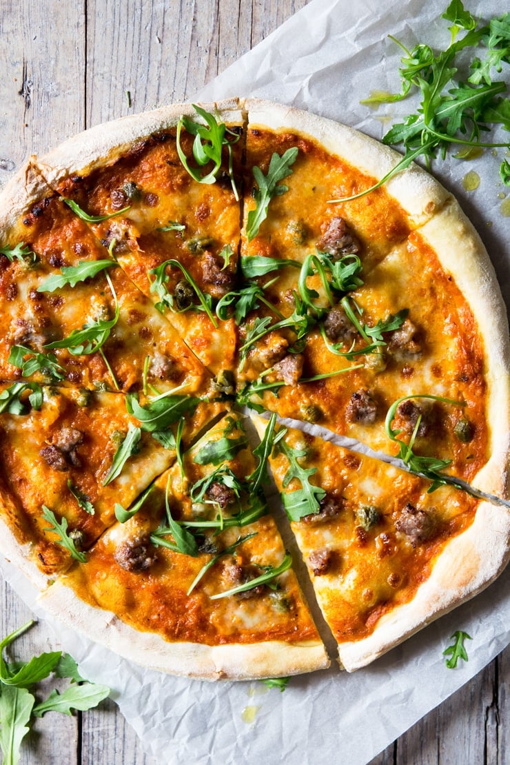 An overhead shot of pizza with sausage, roasted red pepper sauce, mozzarella and arugula sitting on a wooden surface