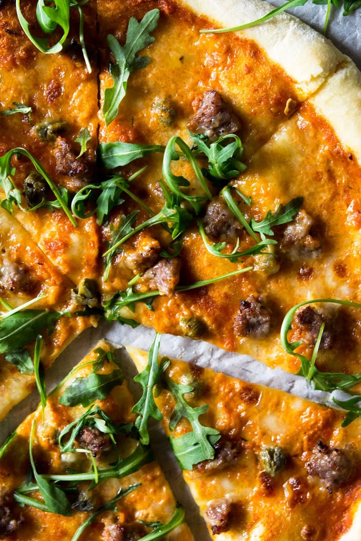 A close up of pizza with sausage and roasted red pepper sauce, melted mozzarella and topped with arugula.