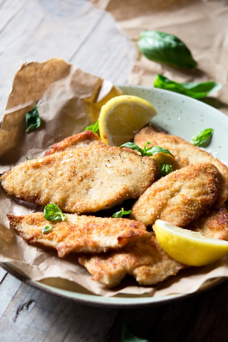 A side shot of breaded chicken cutlets on a plate with lemon wedges