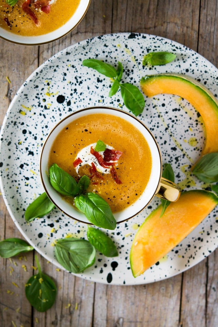 An overhead shot of melon gazpacho soup in a mug sitting on a patterned plate with slices of melon