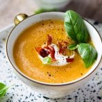 A close up of melon gazpacho in a mug topped with creme fraiche, crispy bacon and basil