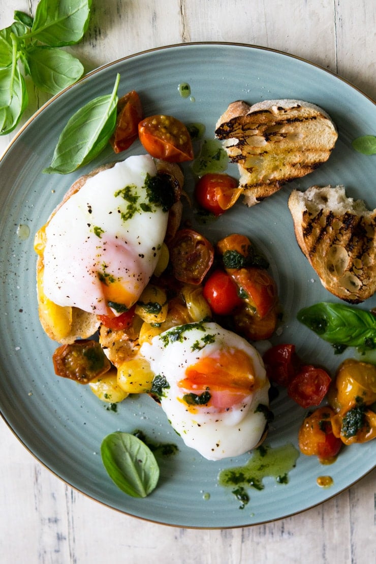 AN overhead shot of sauteed tomato breakfast crostini with poached eggs on top and scattered with basil