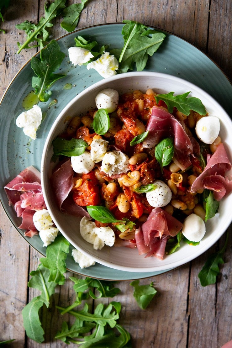 An overhead shot a warm white bean salad in a white bowl topped with prosciutto and mozzarella