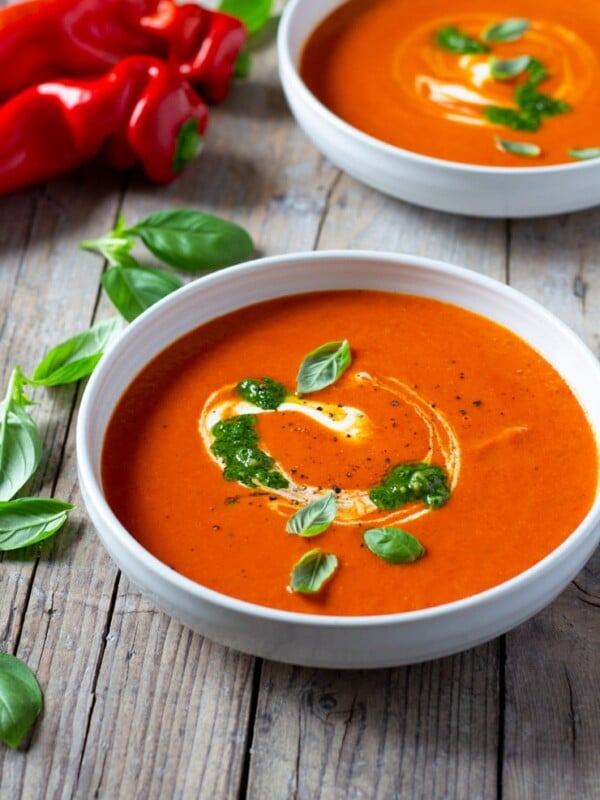 A bowl of roasted red pepper soup on a wooden board with peppers and basil in the background