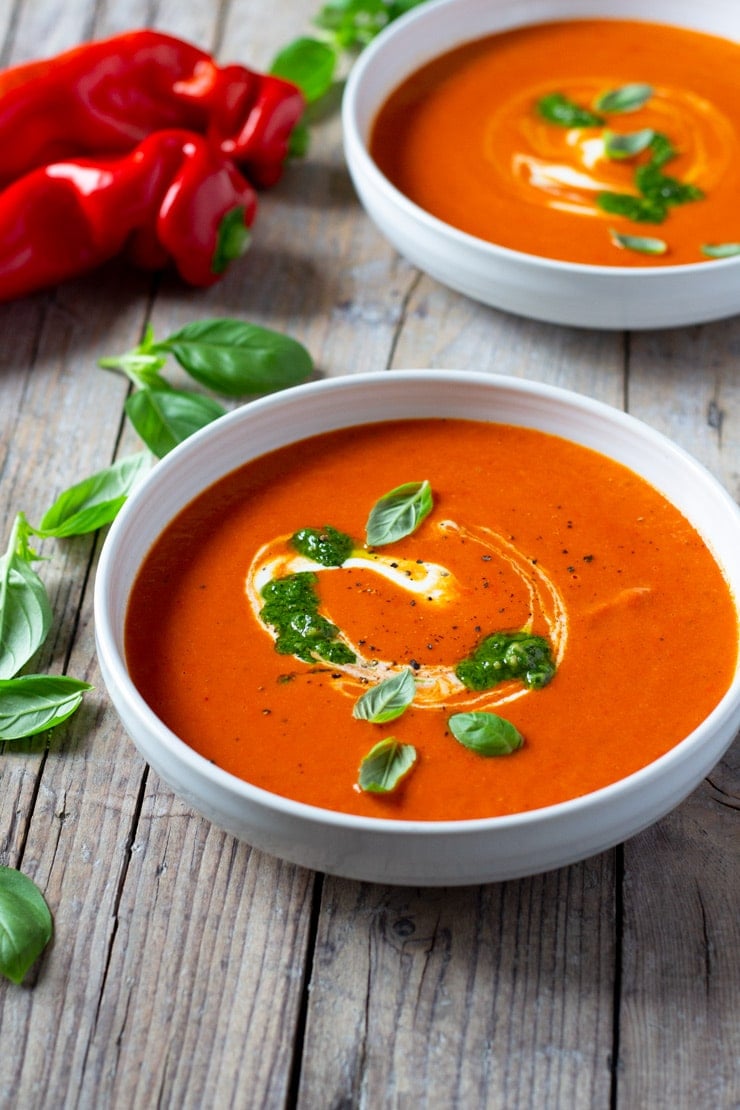 A bowl of roasted red pepper soup on a wooden board with peppers and basil in the background