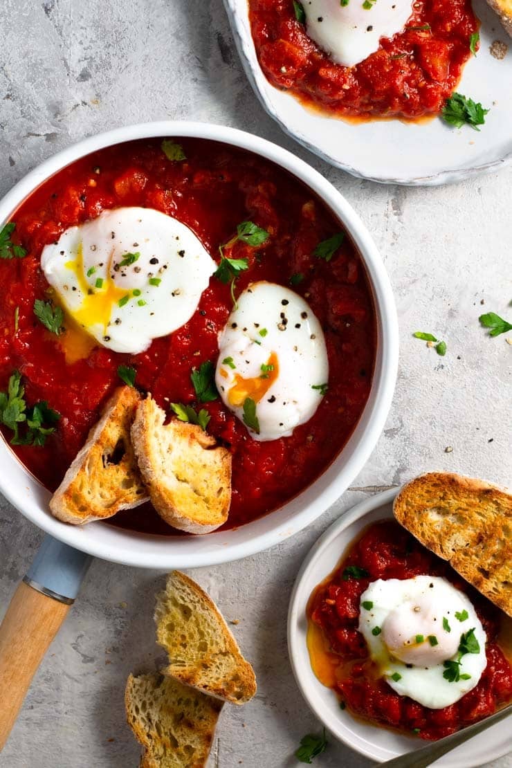 An overhead shot of eggs in purgatory in a frying pan and on two plates with crusty bread