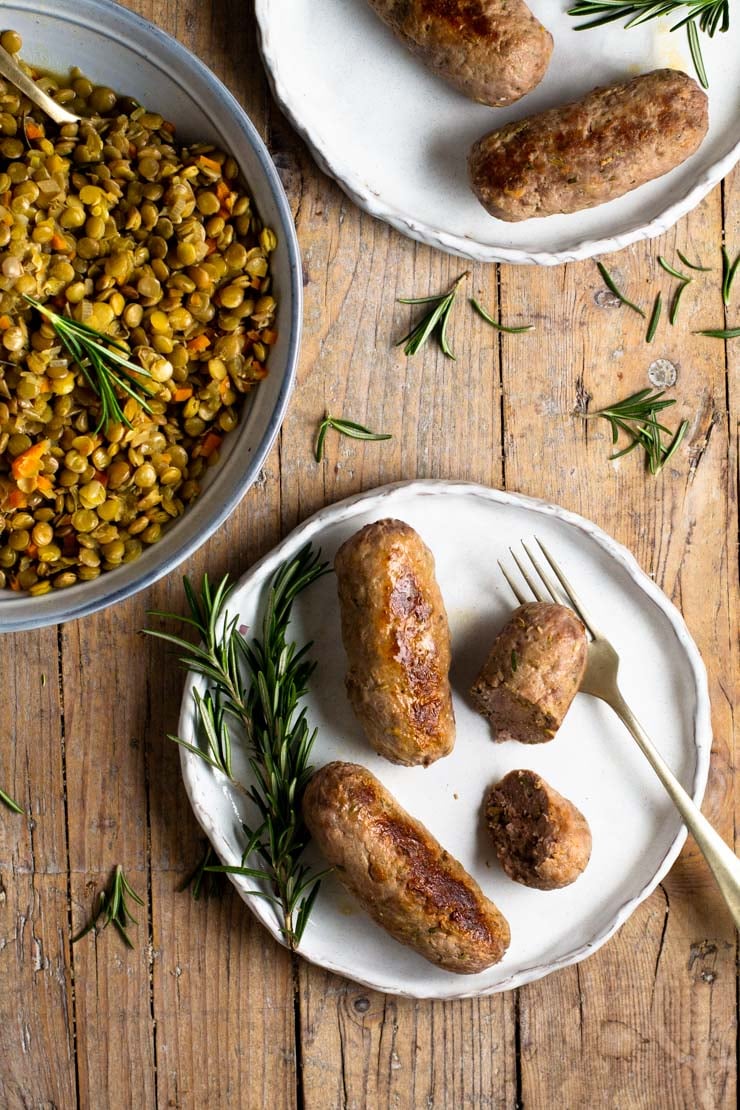 Homemade sausage on a plate with a bowl of lentils in the middle and rosemary scattered around