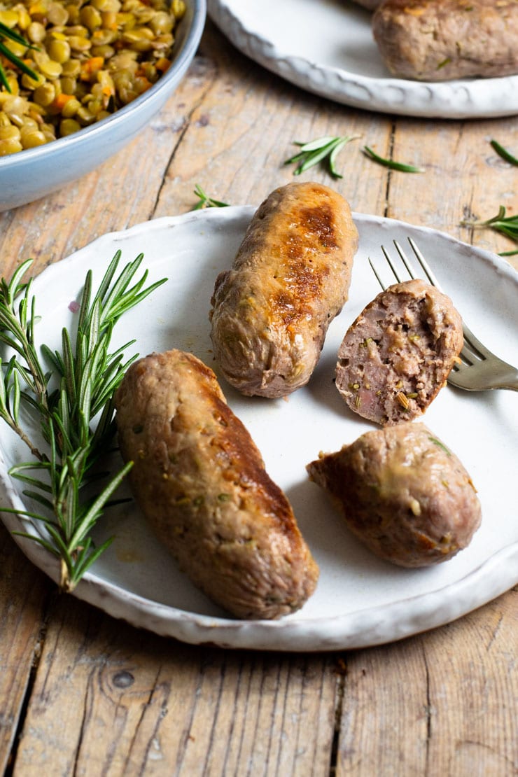 Two whole homemade sausages on a plate and another one cut in half with a rosemary sprig at the side