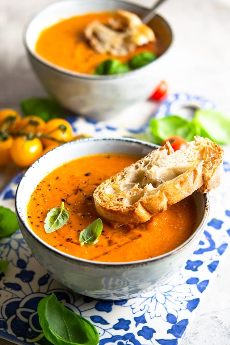 A close up of a bowl of roasted tomato soup with a slice of crusty bread at the side and garnished with basil