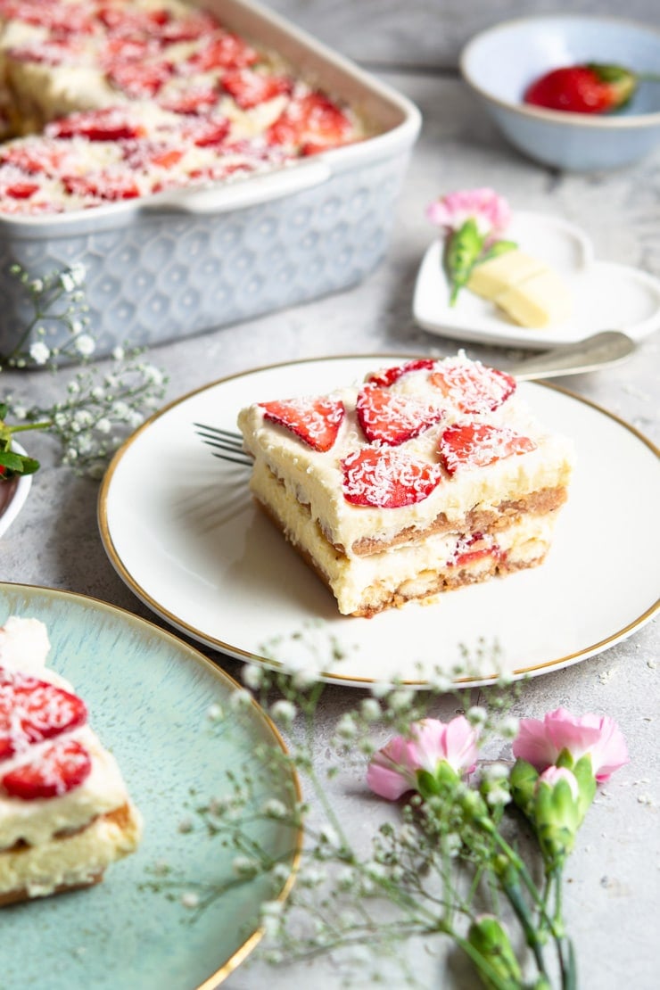 A slice of strawberry tiramisu on a white plate with flowers scattered around 