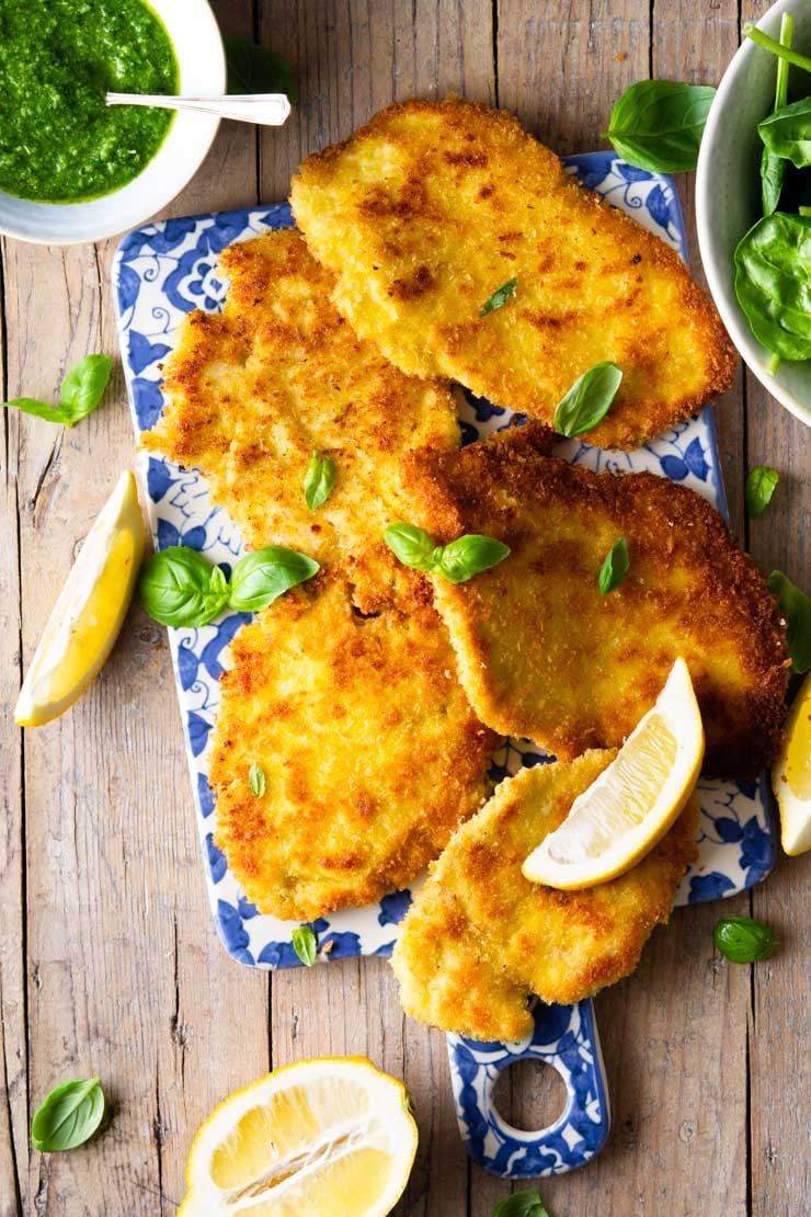 An overhead shot of Italian chicken cutlets on a blue board with lemon wedges and pesto