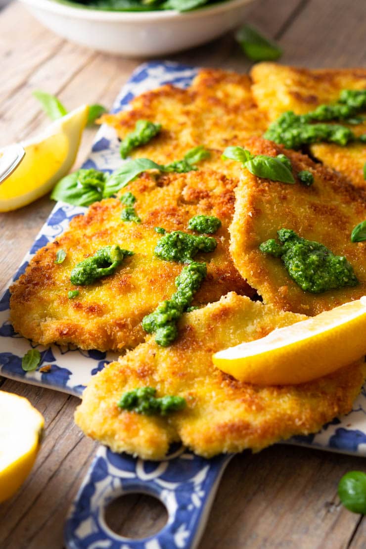 A side shot of breaded chicken cutlets on a blue serving board drizzled with pesto and lemon juice