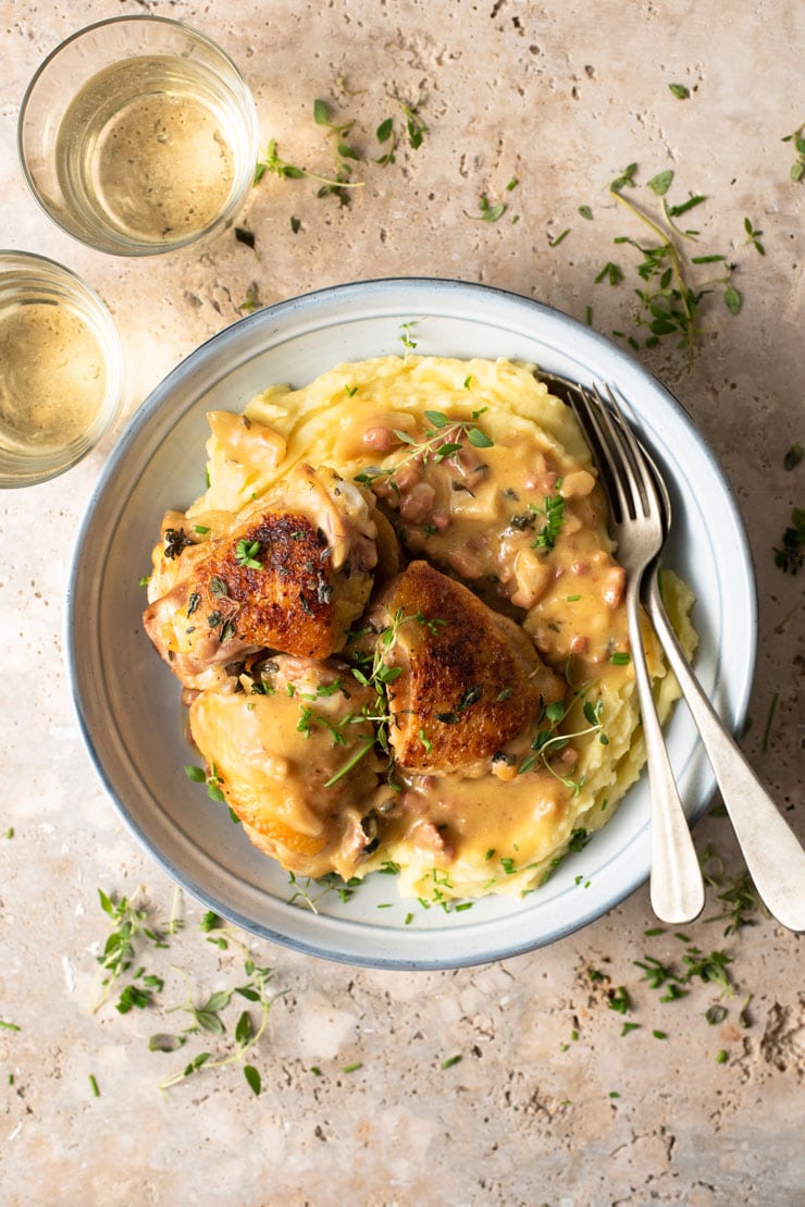 An overhead shot of creamy Tuscan chicken in a blue bowl with mashed potatoes and glasses of white wine