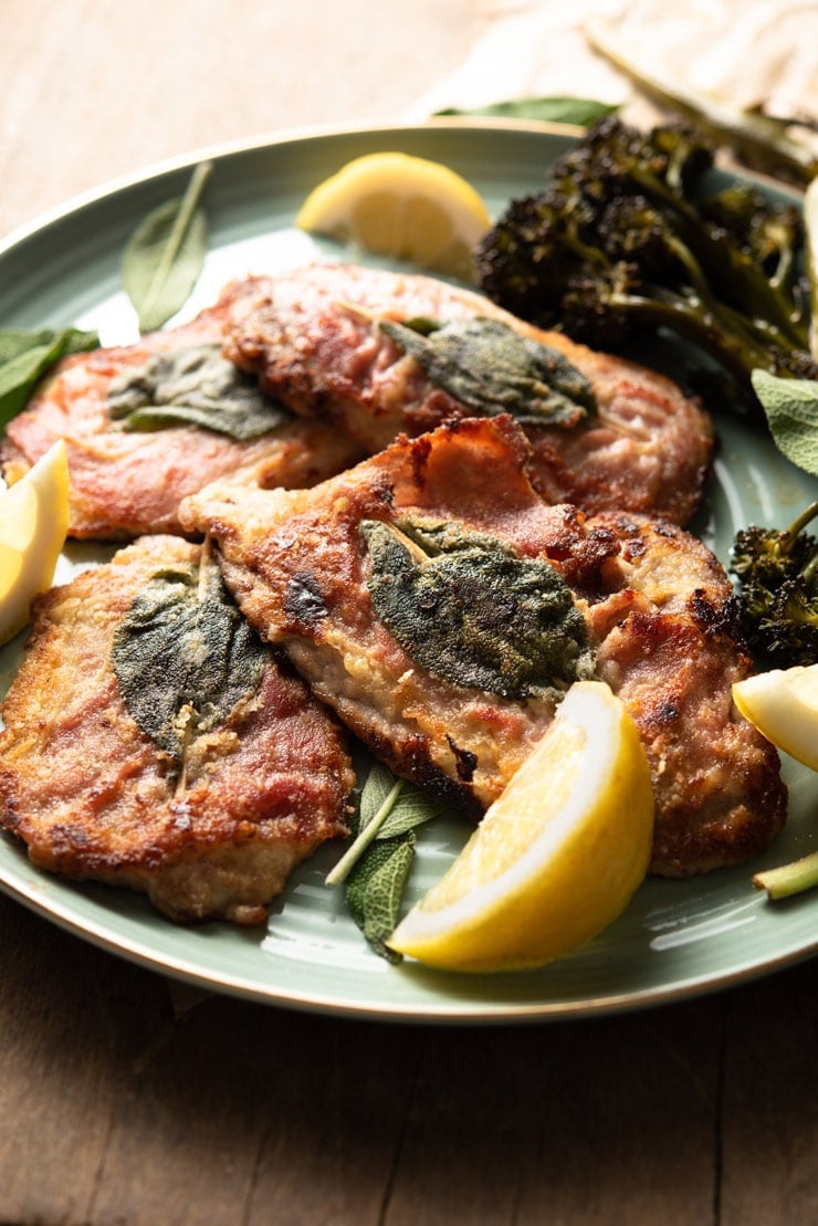 A side shot of veal saltimbocca with prosciutto and sage on a plate with lemon wedges