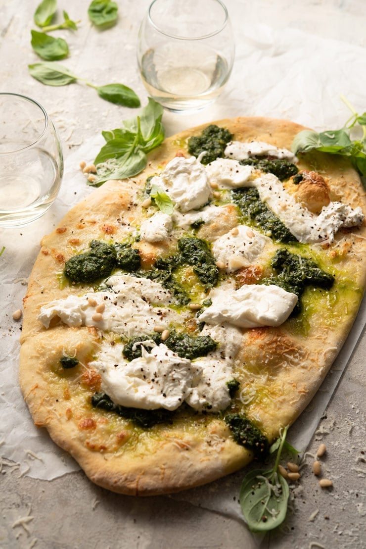 A basil pizza with burrata and pesto on a wooden surface with glasses of white wine in the background