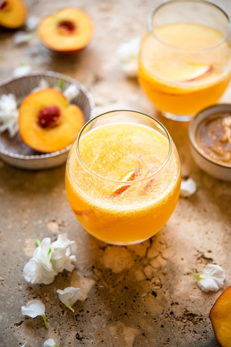 A peach bellini cocktail in a glass with peaches and peach puree in the background