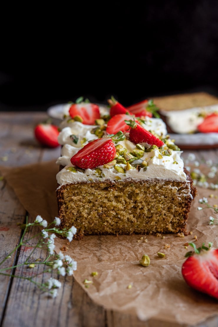 A side shot of a cut pistachio cake sitting on a wooden surface with strawberries and cream