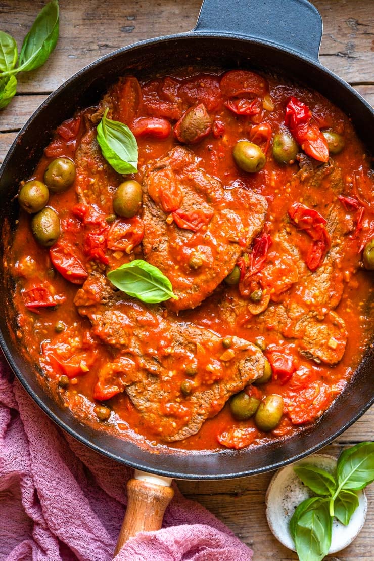 Overhead shot of a skillet with steak in a tomato sauce with olives and basil