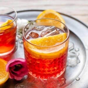 A close up of a Negroni cocktail on a pewter plate garnished with orange slices and a rose sitting beside it
