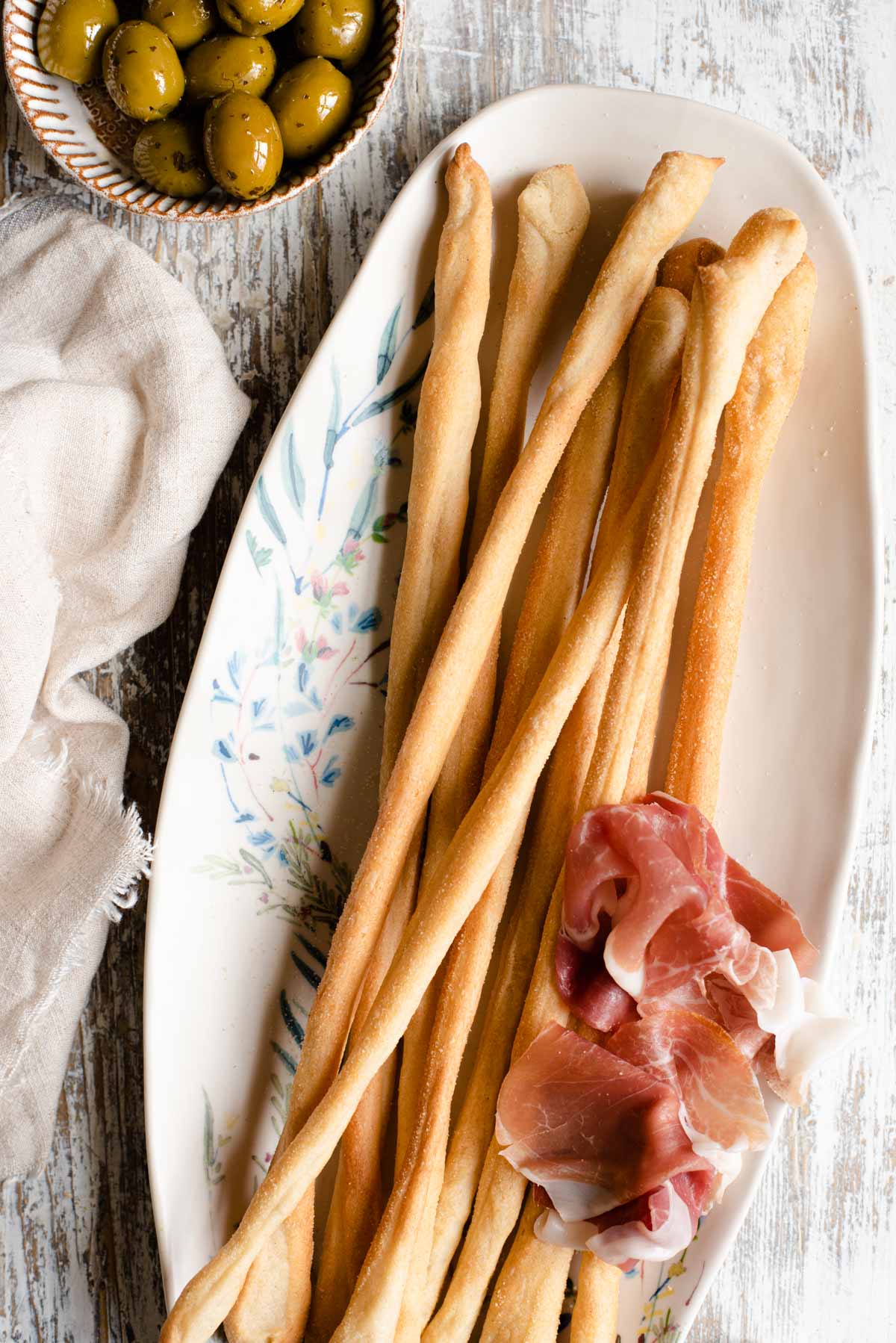 An overhead shot of Italian breadsticks on a serving plate with olives and prosciutto