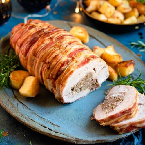 A stuffed turkey breast on a serving plate with slices cut and roasted potatoes