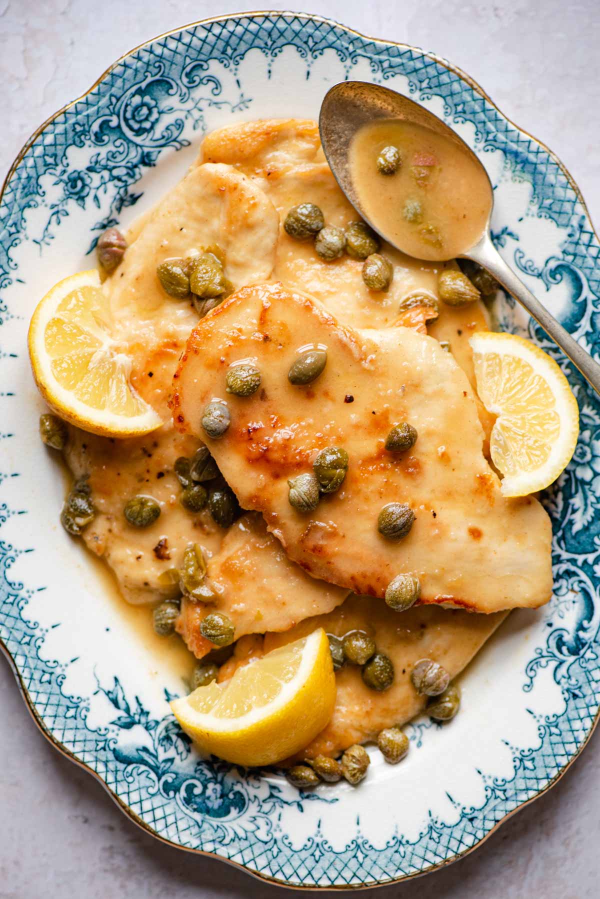 An overhead shot of chicken piccata on a blue serving plate with lemon wedges and capers