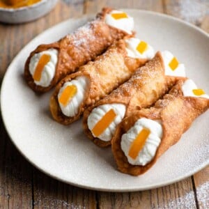 Four cannoli on a plate filled with ricotta and a slice of candied orange