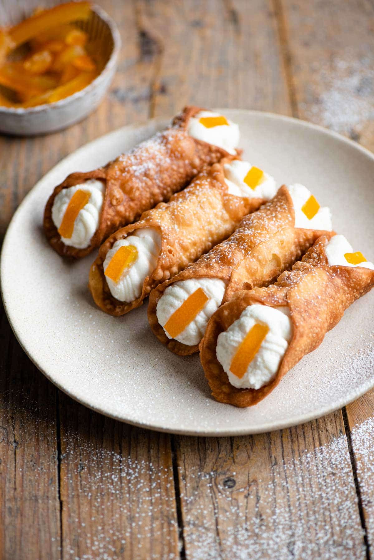 Four cannoli on a plate filled with ricotta and a slice of candied orange