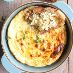 A close up cropped square image of baked ricotta in a blue ceramic round dish topped with a slice of crusty bread dipped in