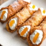A close up of four homemade cannoli filled with ricotta and topped with candied orange on a plate