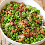 A close up of peas and pancetta in an oval serving dish on a wooden surface