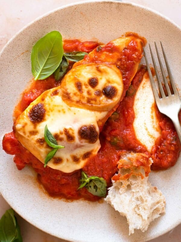 A close up of a piece of chicken pizzaola on a plate with crusty bread and basil leaves