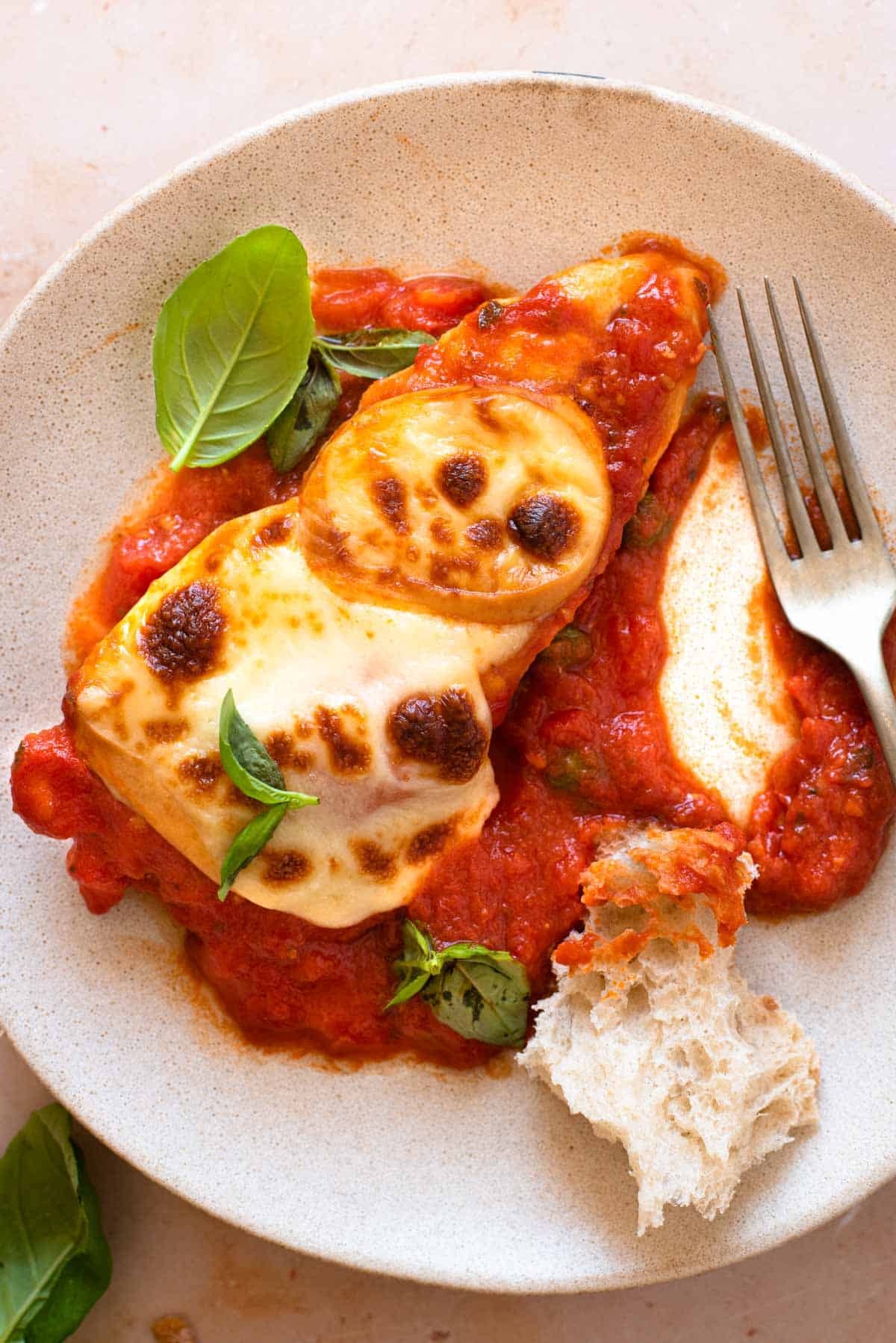 A close up of a piece of chicken pizzaola on a plate with crusty bread and basil leaves