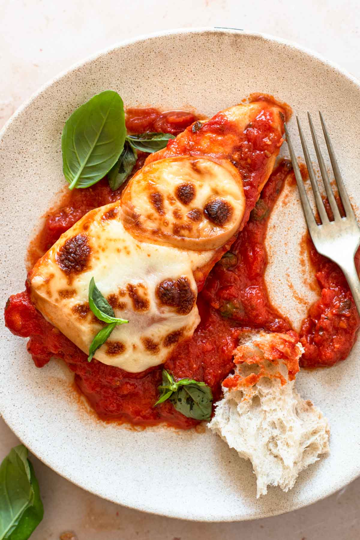 A close up of a piece of chicken pizzaola on a plate with crusty bread and basil leaves